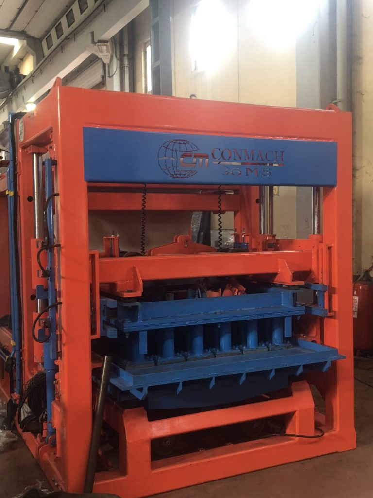 Turuncu gövdeli ve mavi detaylı "CONMACH 36 M5" marka beton blok makinesinin iç kısmında yer alan çoklu kalıp sistemi görülmektedir. Makine, endüstriyel bir üretim tesisinin içinde konumlandırılmış olup sağlam metal şasisi ve hidrolik aksamlarıyla dikkat çekmektedir.