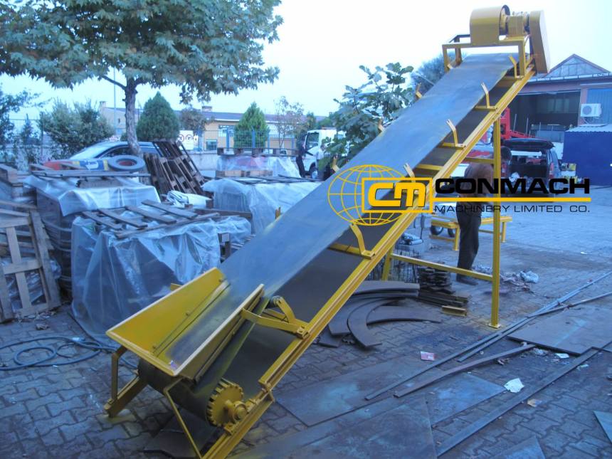 Beton santrallerinde ham madde transferi için kullanılan, sarı boyalı, eğimli metal gövdeli konveyör bant sistemi. Üzerinde Conmach logo filigranı görülmektedir.