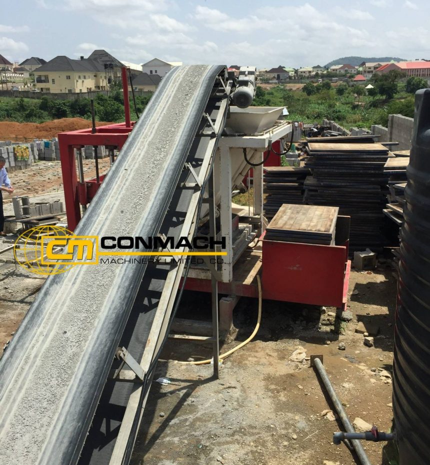 Açık havada kurulu bir tesiste, beton agregasını ana makineye taşıyan eğimli siyah bir konveyör bant ve arkada istiflenmiş ahşap üretim paletleri görülmektedir. Makine üzerinde sarı-siyah logolu "CONMACH" yazısı yer almakta, arka planda ise yerleşim alanı ve yeşil bir arazi uzanmaktadır.