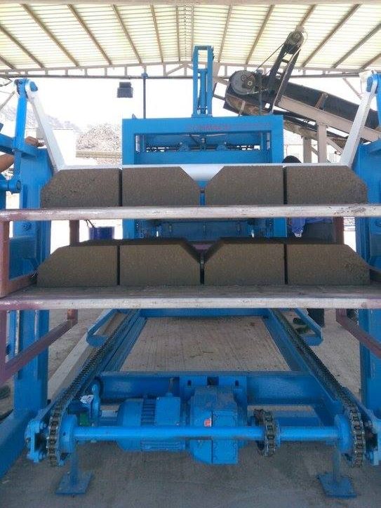 Mavi renkli bir makinenin çıkış bandında, metal paletler üzerinde iki sıra halinde dizilmiş taze beton bloklar yer almaktadır. Blokların üst kısımları eğimli bir tasarıma sahip olup kuruma aşamasına geçmeye hazırdır.